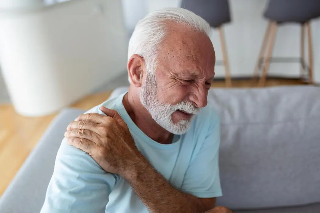 Older man on a couch holding his painful shoulder and neck